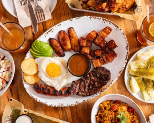 A top-down view of a large plate featuring grilled steak, crispy pork belly cubes, fried plantains, sliced avocado, arepas, a sunny-side-up egg over rice, and a grilled sausage, served with a cup of brown sauce. Surrounding dishes include coleslaw, yuca with green sauce, yellow rice with vegetables, and several glasses of iced tea. The food is arranged on a wooden table in a colorful, inviting presentation.