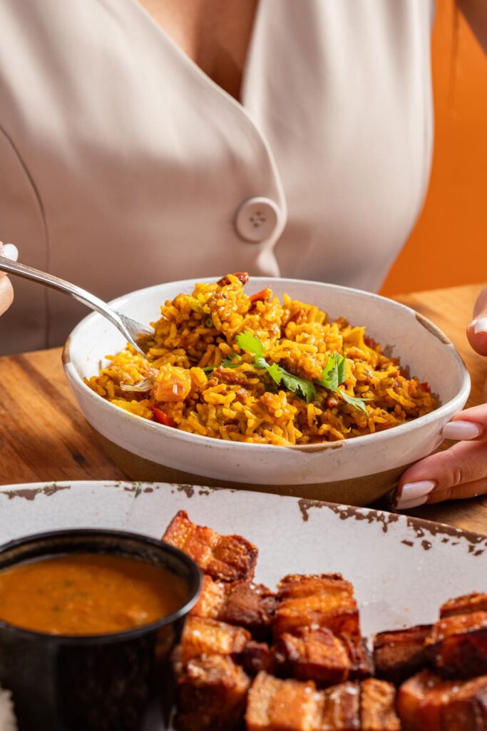 Close-up of a person holding a bowl of seasoned yellow rice with vegetables and cilantro, with a plate of crispy pork chunks and sauce partially visible in the foreground.