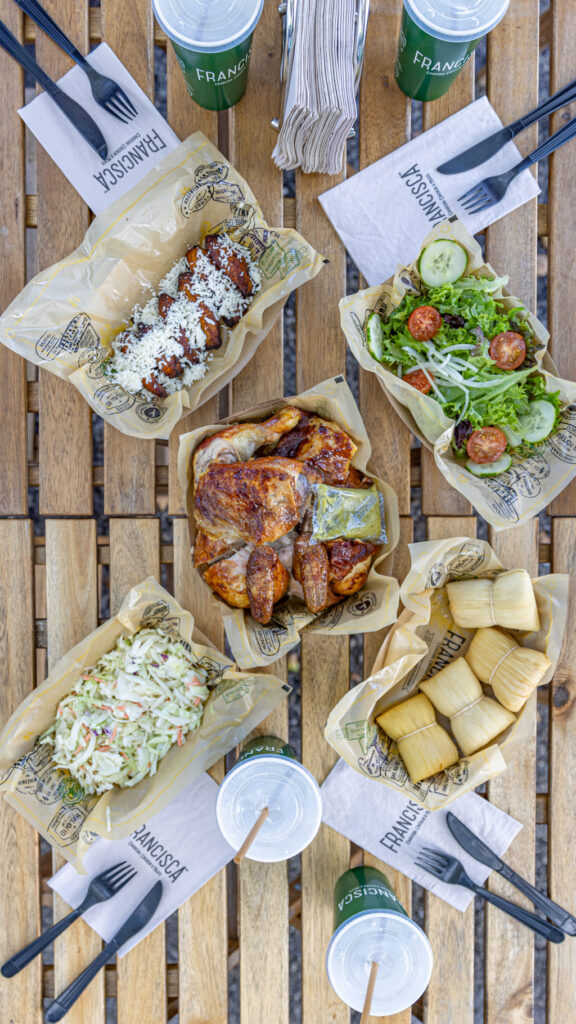 Top-down view of a wooden outdoor table filled with food trays from a restaurant. The spread includes roasted chicken with a packet of green sauce, a tray of grilled plantains topped with white cheese, a fresh green salad with cucumbers and cherry tomatoes, a tray of small wrapped corn cakes, and a serving of creamy coleslaw. Several green branded drink cups with lids and straws, napkins, and black plastic utensils are arranged around the table.