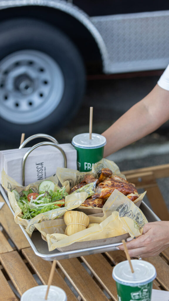 A person sets down a metal tray filled with food, including roasted chicken pieces, a fresh green salad with cucumbers and tomatoes, and several small wrapped corn cakes. A green disposable cup with a lid and straw sits on the tray. The scene takes place outdoors on a wooden table, with part of a food truck wheel visible in the background.