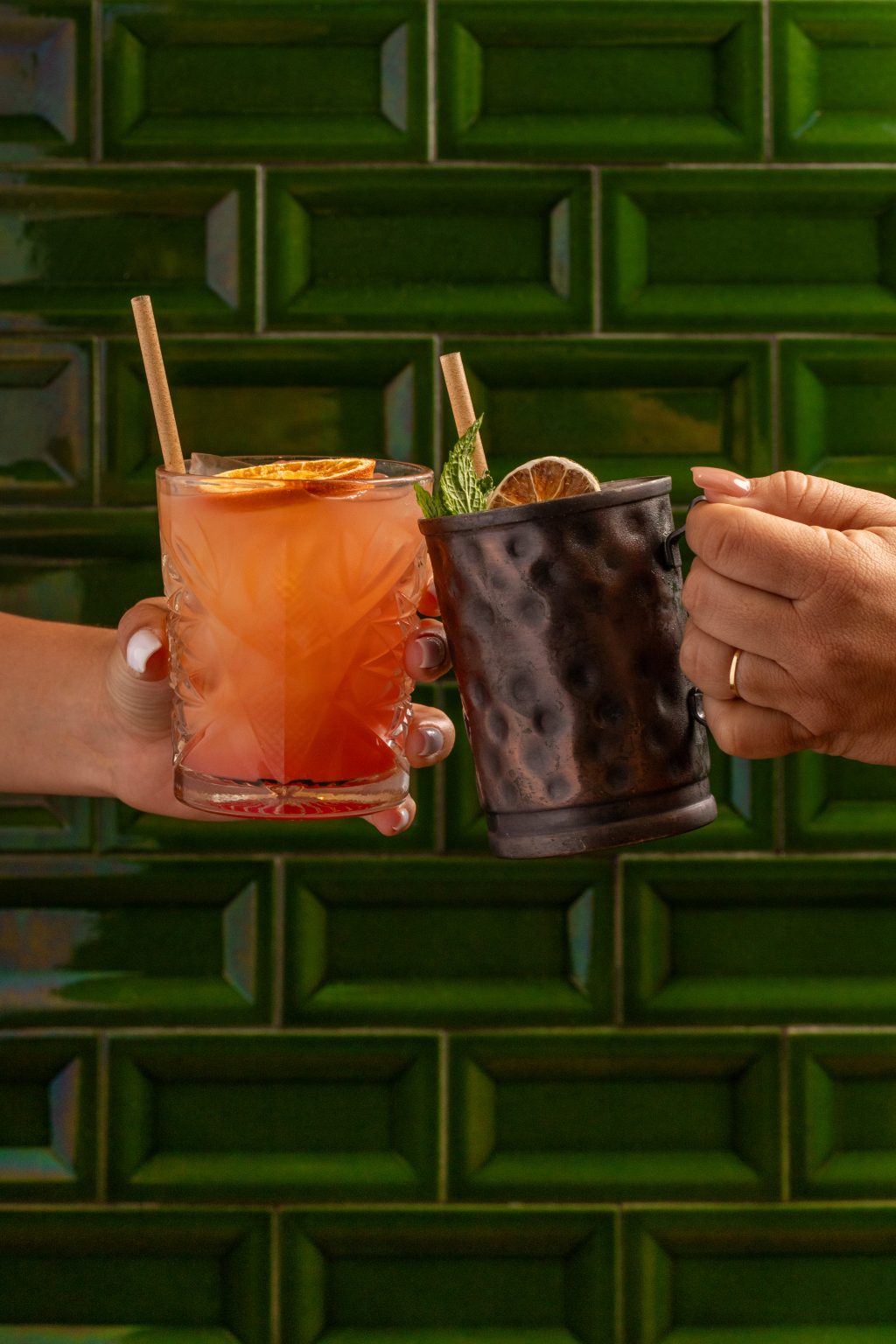 Two hands clink colorful cocktails: one in a crystal glass with citrus slices, the other in a dark hammered metal mug garnished with mint, against a green tiled background.