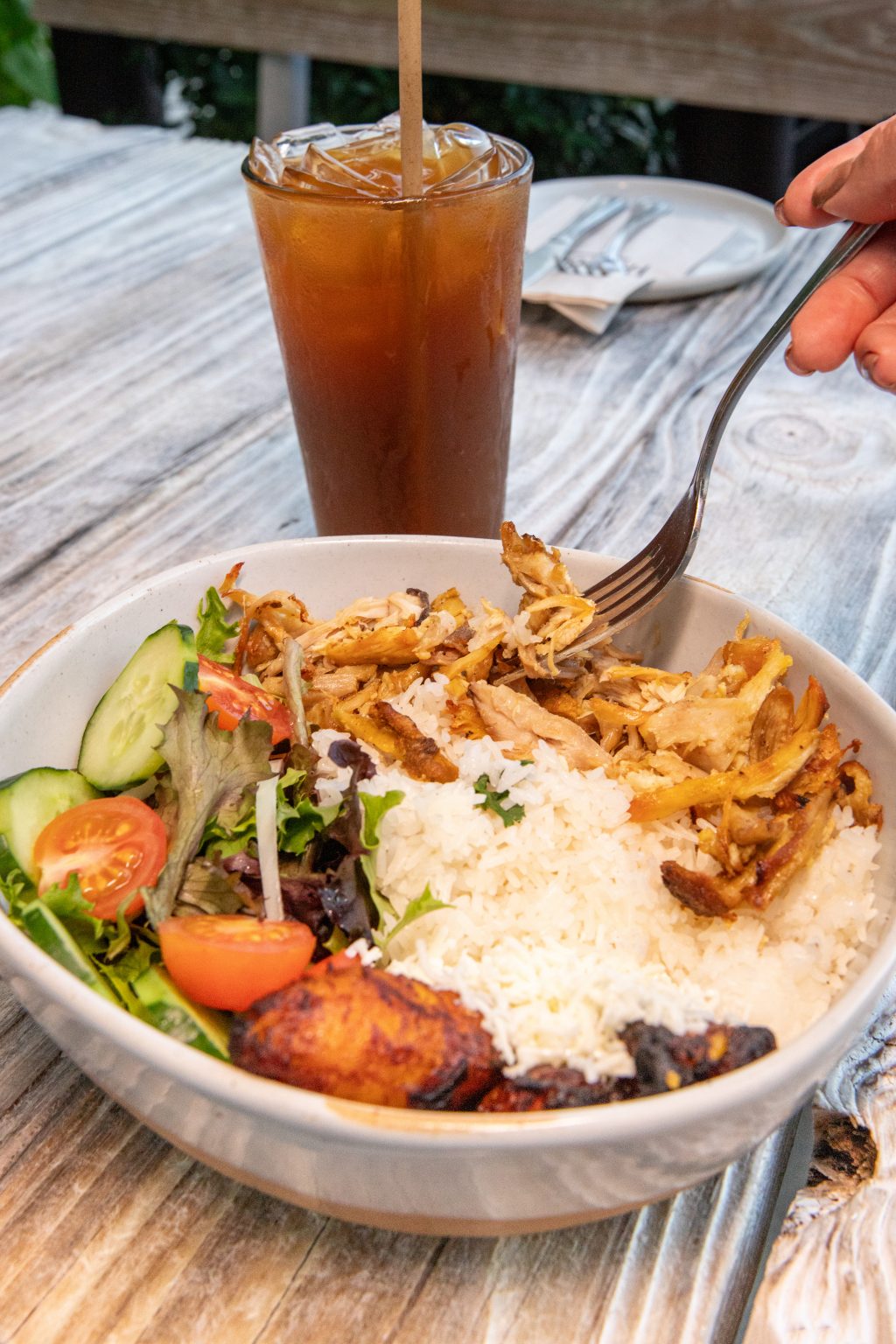 A bowl filled with shredded roasted chicken, white rice, fresh salad with cucumbers and cherry tomatoes, and fried plantains, with a person lifting a forkful of chicken. A tall glass of iced tea sits behind the bowl on a rustic wooden table.