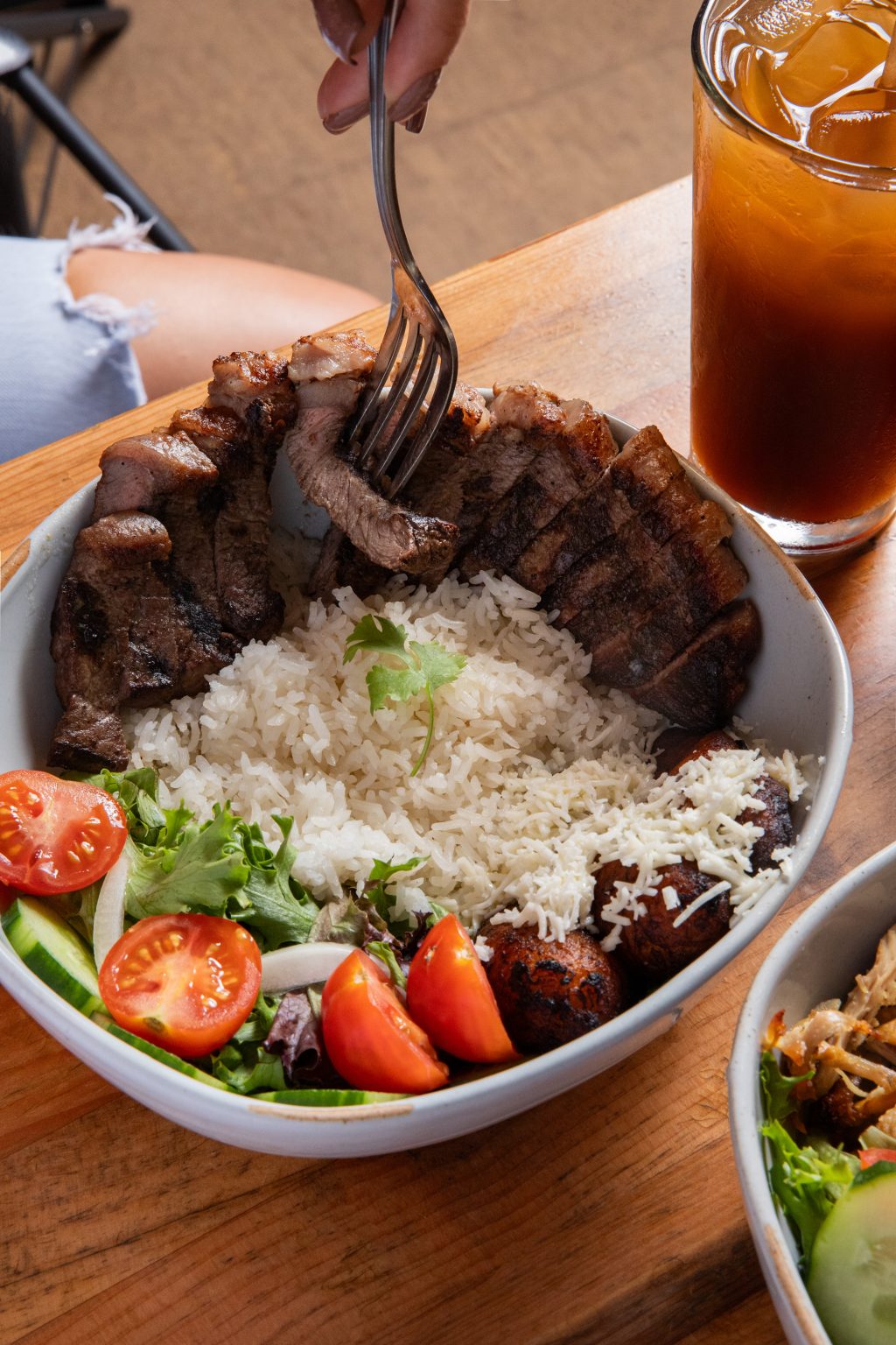 A bowl filled with sliced grilled beef served over white rice, accompanied by fried plantains topped with cheese, mixed greens, cucumber slices, and cherry tomatoes. A person is cutting into the beef with a fork, and a glass of iced tea sits beside the bowl on a wooden table.