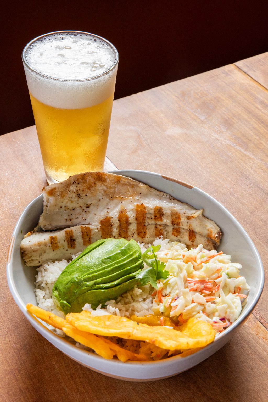 A bowl of food featuring grilled fish fillets over white rice, sliced avocado, creamy coleslaw, and fried plantain strips, placed on a wooden table. Behind the bowl sits a tall glass of cold beer with a frothy head.