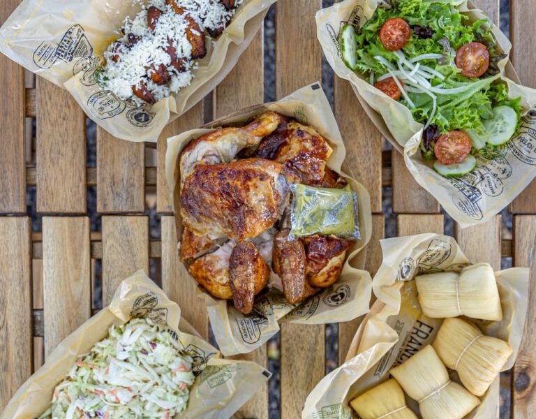 Overhead view of a table filled with takeout dishes, including roasted chicken, fresh salad, coleslaw, cheese-topped fried appetizers, and wrapped tamales, along with drinks and wrapped utensils on a wooden outdoor table.