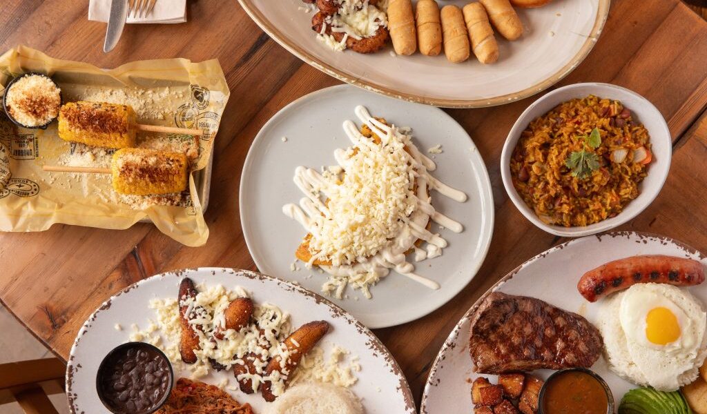 A top-down view of several Venezuelan and Latin-inspired dishes, including grilled corn with cheese, fried plantains topped with cheese, tequeños, empanadas, a bowl of yellow rice with vegetables, shredded beef with black beans and rice, and a plate with grilled steak, fried pork bites, sausage, avocado slices, and a sunny-side-up egg.