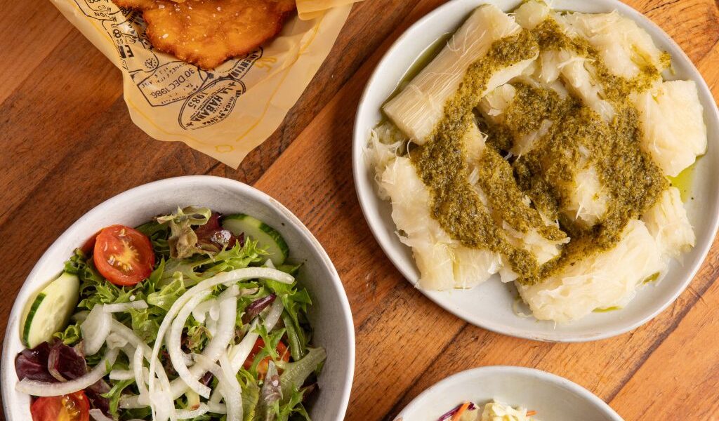 A top-down view of assorted side dishes including tostones, sweet plantains with cheese, yuca with green sauce, a fresh garden salad with tomatoes and cucumbers, coleslaw, and several wrapped hallacas on a wooden table.