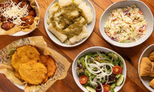 A top-down view of assorted side dishes including tostones, sweet plantains with cheese, yuca with green sauce, a fresh garden salad with tomatoes and cucumbers, coleslaw, and several wrapped hallacas on a wooden table.