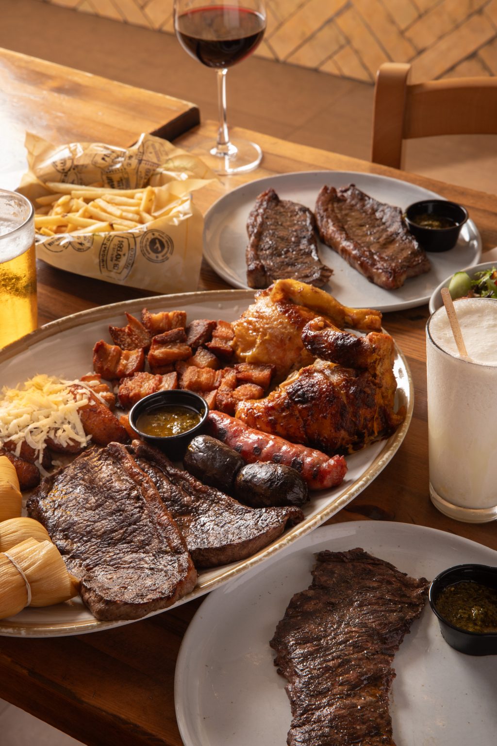 A table filled with assorted grilled meats including steak, chicken, sausages, and chicharrón served on large platters, accompanied by fries, dipping sauces, a glass of beer, a glass of red wine, and a creamy beverage.