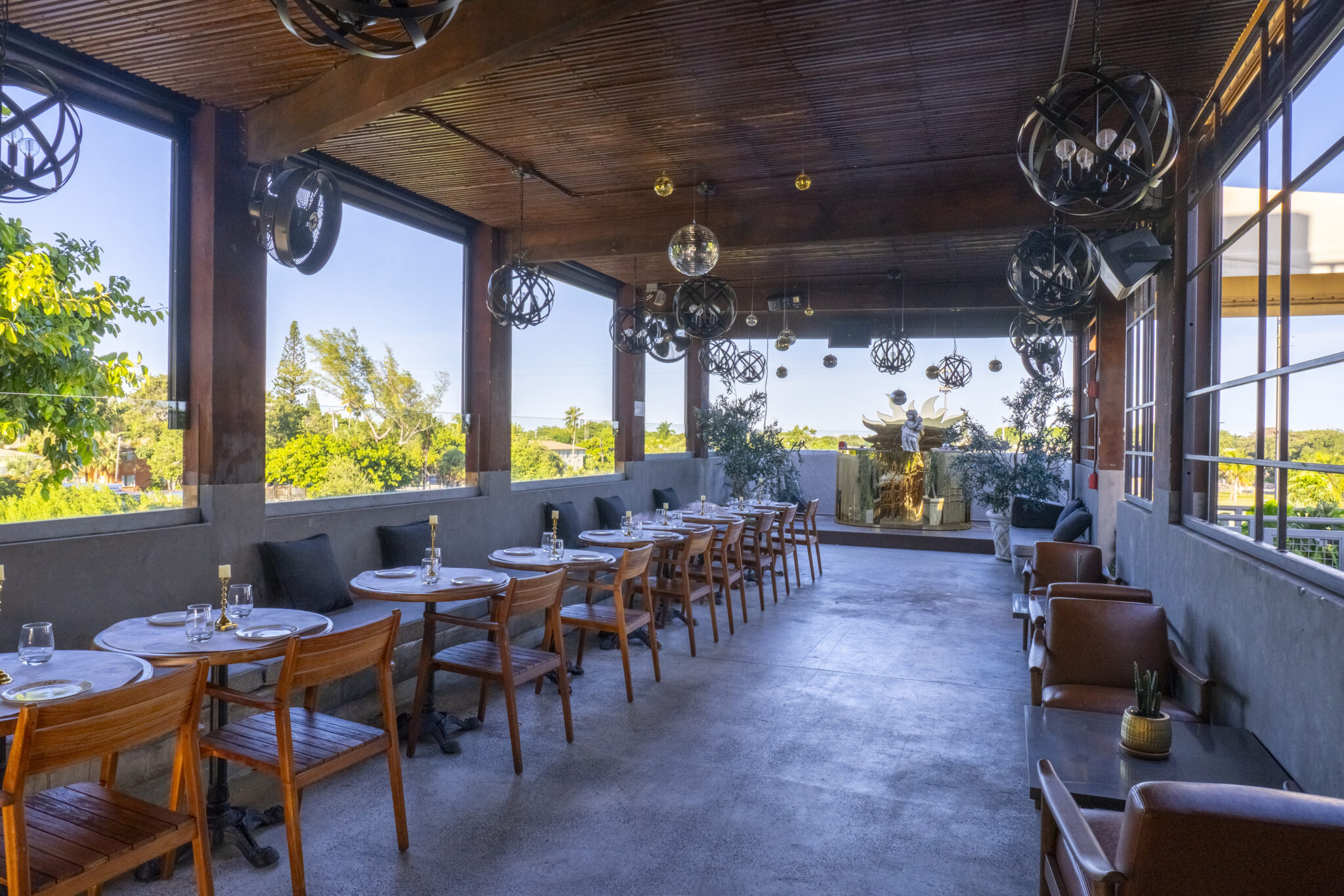 Open-air restaurant terrace with wooden ceiling, modern spherical light fixtures, and rows of wooden tables and chairs overlooking lush green treetops. A decorative golden sculpture sits at the far end, with lounge seating arranged along the right side.