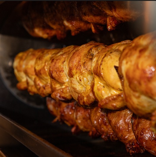 Close-up of whole chickens roasting on a rotisserie, with golden-brown skin glistening as they cook.