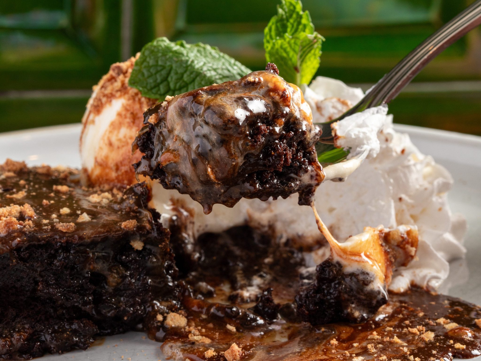 Close-up of a gooey chocolate brownie with melted marshmallow, caramel, and whipped cream, lifted by a fork with a mint garnish in the background.