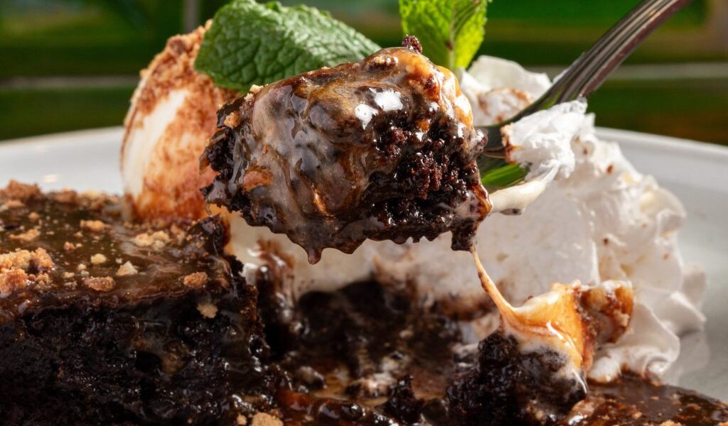 Close-up of a gooey chocolate brownie with melted marshmallow, caramel, and whipped cream, lifted by a fork with a mint garnish in the background.