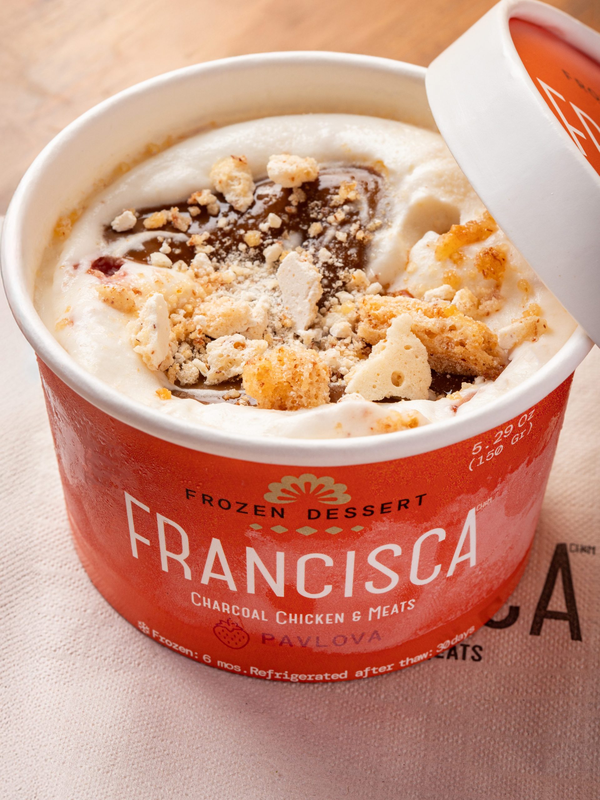 Close-up of a Francisca-branded pavlova frozen dessert cup with its lid slightly open, revealing creamy filling topped with crumbled meringue and caramel swirls.