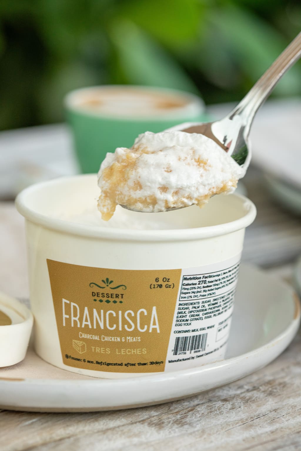 Close-up of a spoon lifting a creamy bite of tres leches dessert from a Francisca-branded cup, with a blurred coffee cup in the background.