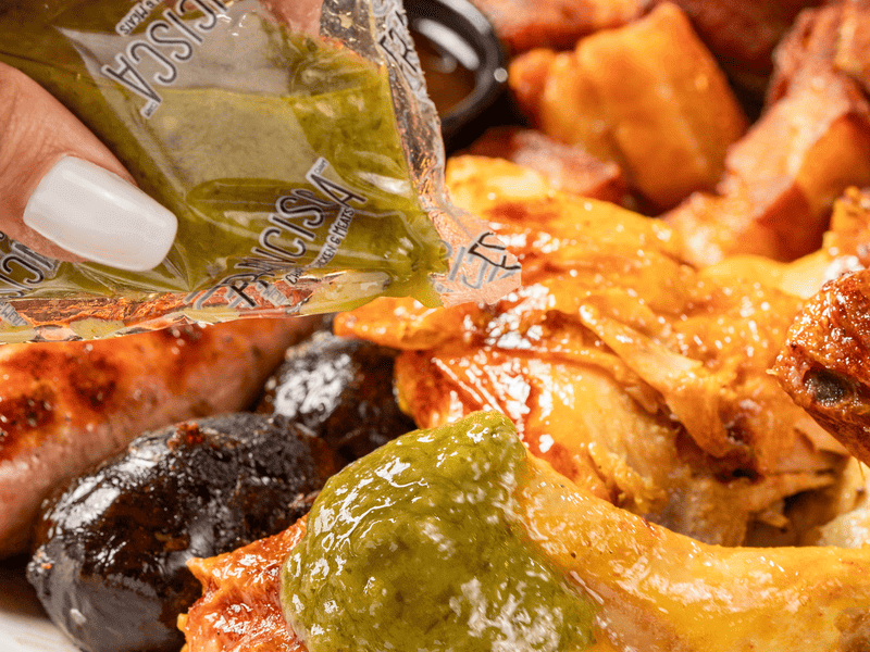 A close-up of a hand squeezing a packet of green sauce over a plate of charcoal-roasted chicken and assorted meats, with crispy sides visible in the background.