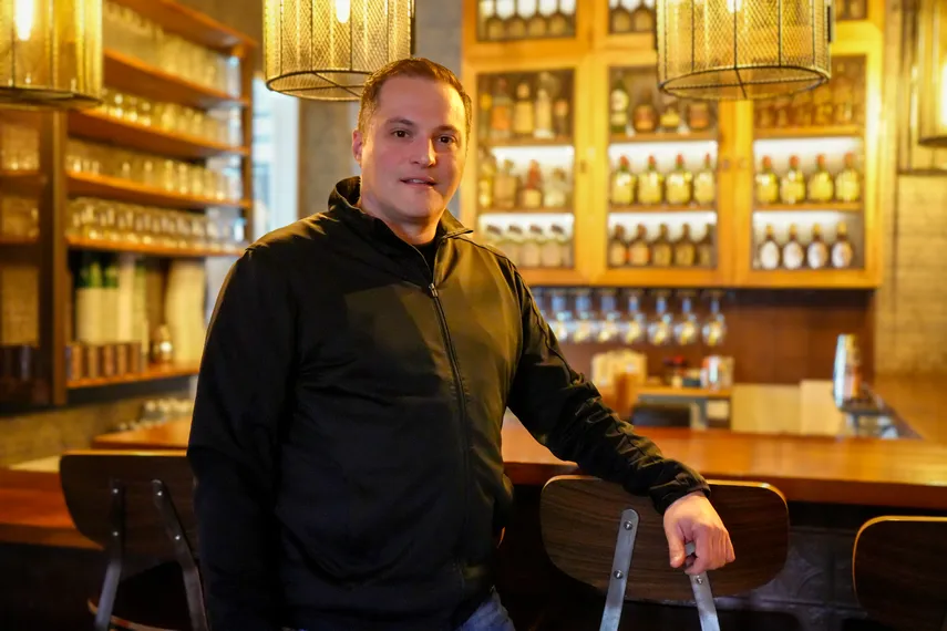 Francisca Restaurant's CEO standing inside a warmly lit bar or restaurant, resting one hand on the back of a wooden barstool. He is wearing a black jacket and looking toward the camera. Behind him are shelves filled with glasses and bottles, along with hanging pendant lights that add a cozy atmosphere.