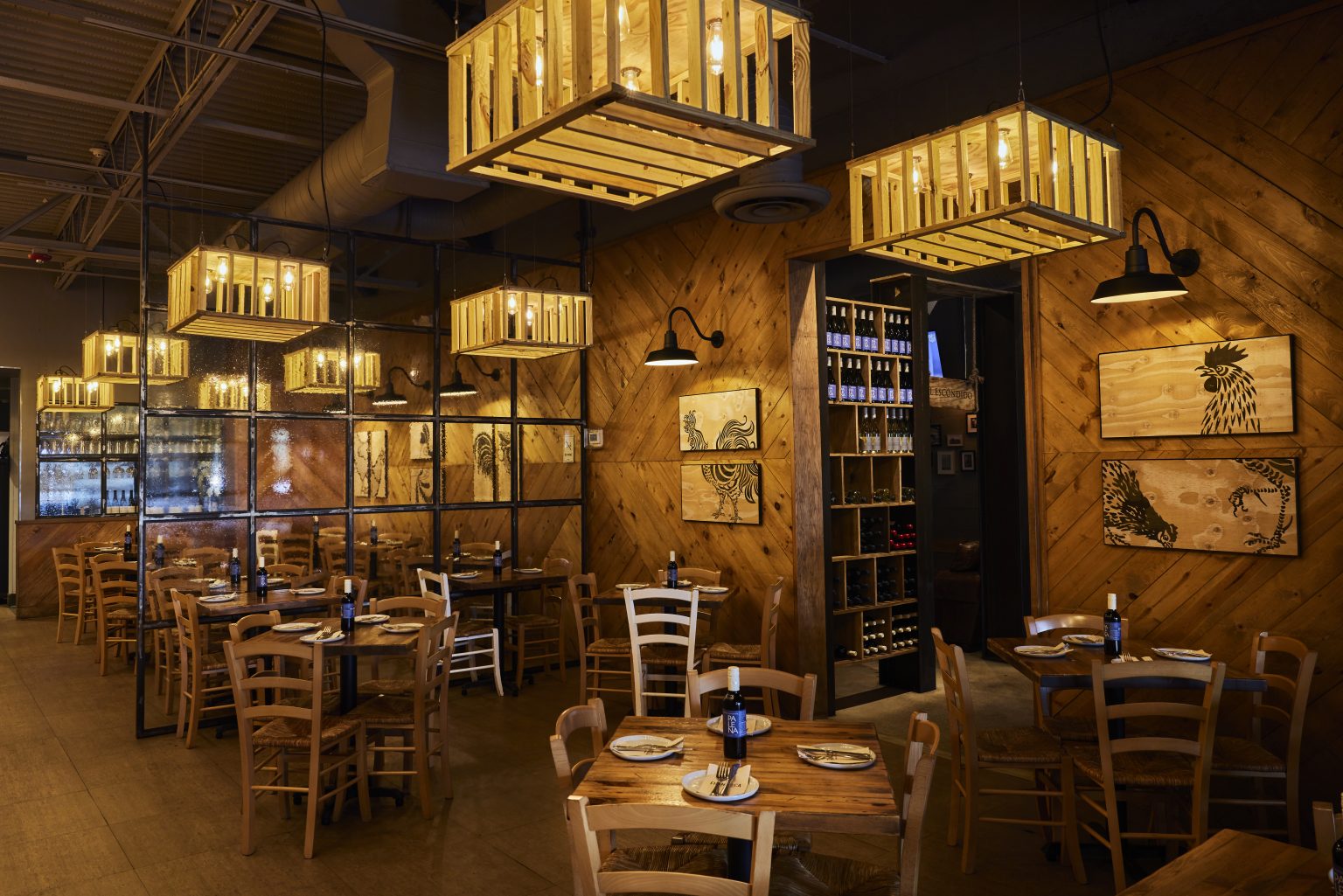 Warm, rustic restaurant interior featuring wooden tables and chairs, crate-style pendant lights, and wood-paneled walls decorated with rooster artwork. A glass partition separates the dining areas, and a wine shelf is visible in the back, creating a cozy, intimate atmosphere.