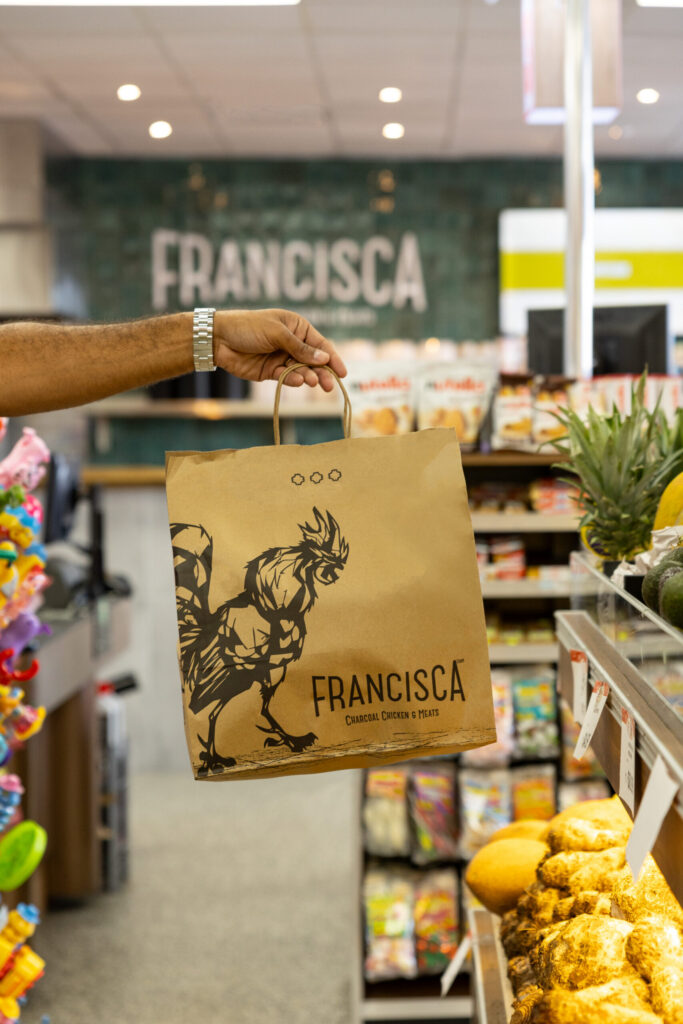 Hand holding a Francisca takeout bag with rooster logo inside a market aisle.