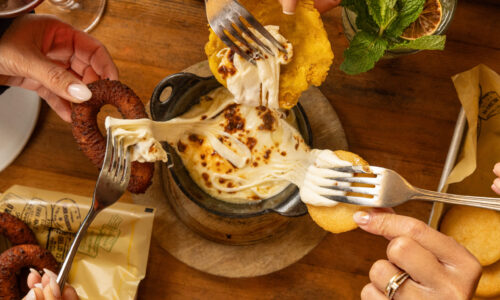 Hands dipping fried items into a skillet of melted, browned cheese on a wooden table.