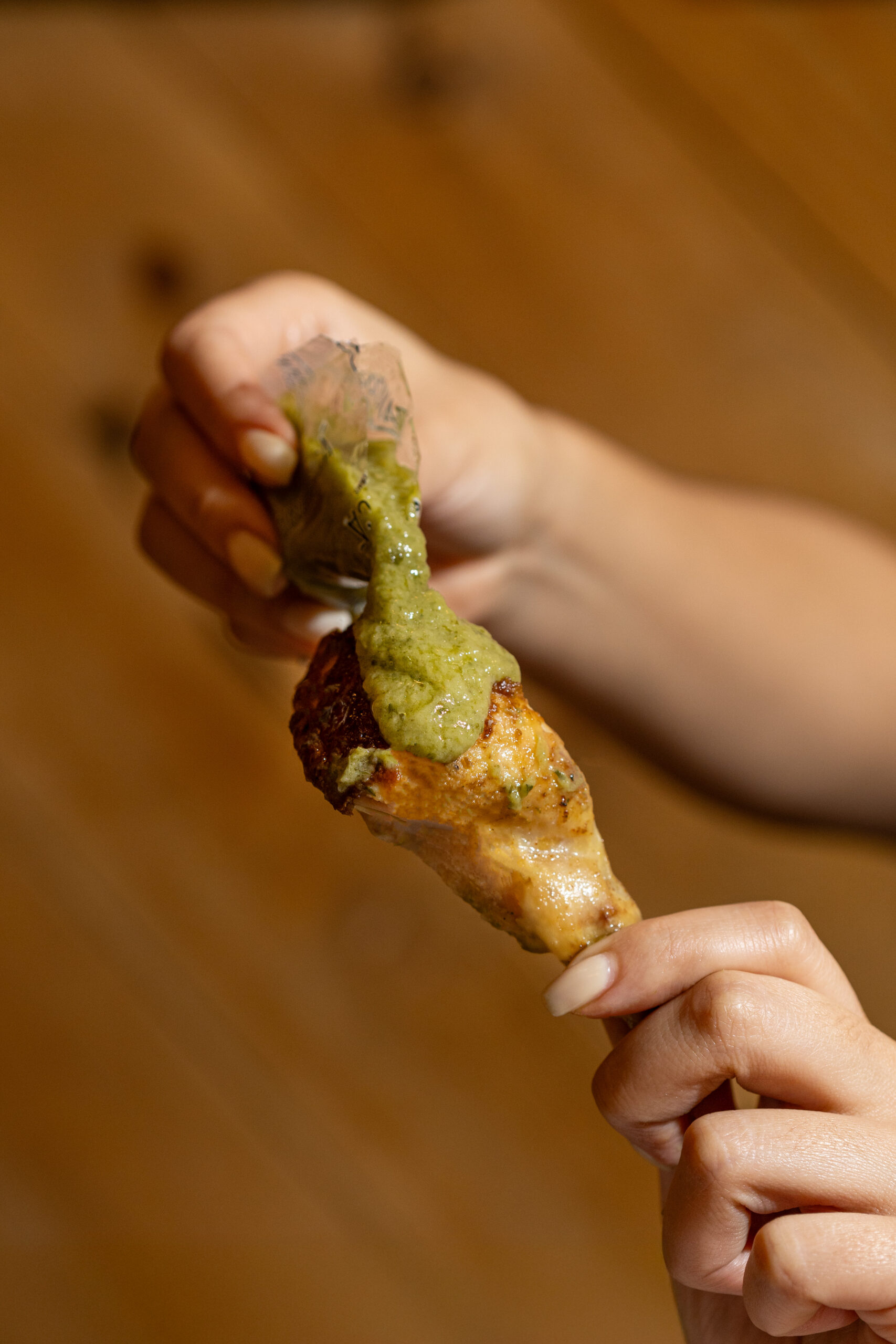 Hand holding a chicken drumstick topped with green sauce over a wooden background.