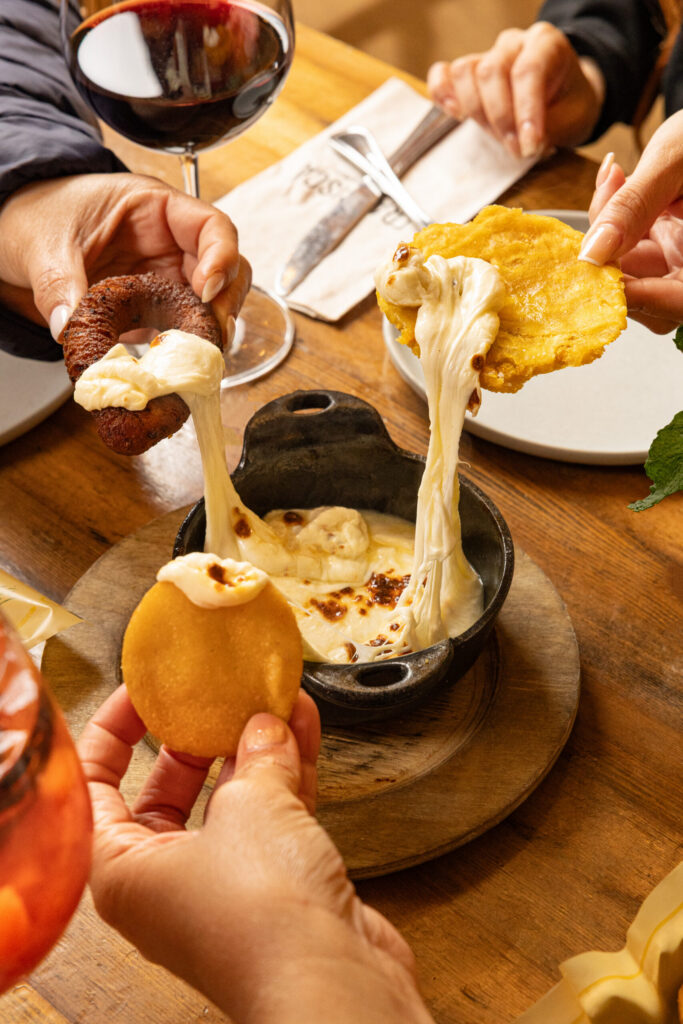 People dipping fried plantains and sausage into melted cheese, creating long cheese pulls over a skillet on a restaurant table