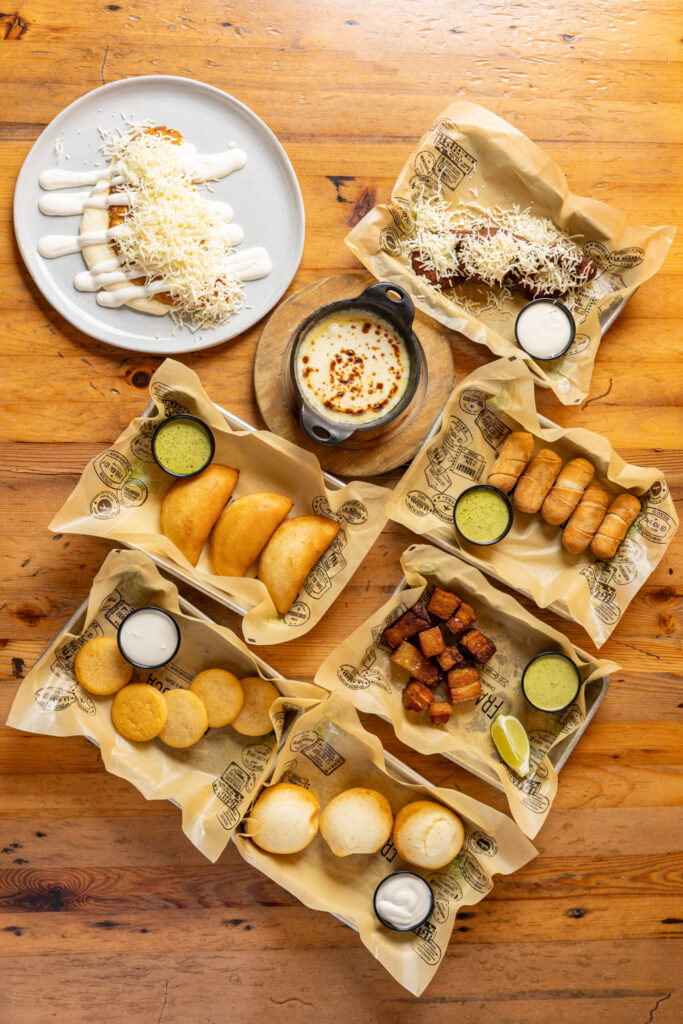 Overhead view of assorted Latin appetizers on a wooden table, including empanadas, cheese bread, fried pork bites with lime, croquettes, melted cheese dip, and desserts topped with shredded cheese and cream sauces.