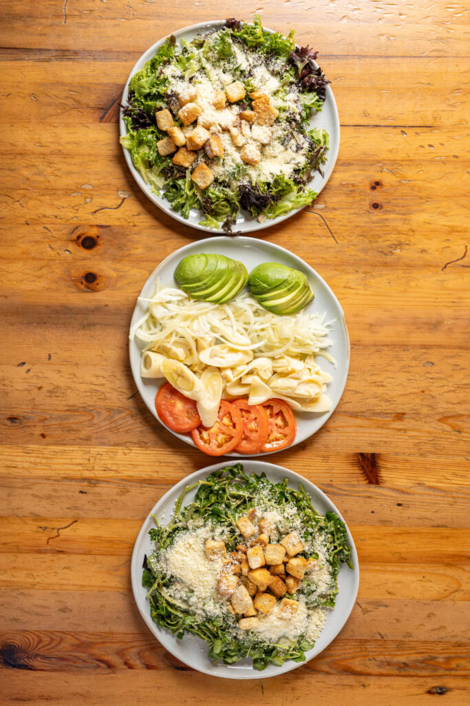 Overhead view of three plates on a wooden table, including Caesar-style salads with croutons and grated cheese, and a plate with sliced avocado, onions, hearts of palm, and tomatoes.