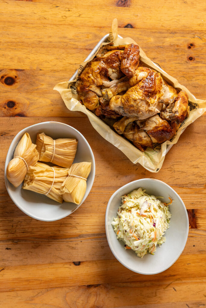 Overhead view of roasted chicken pieces served in a tray with tamales wrapped in corn husks and a side bowl of coleslaw on a wooden table.