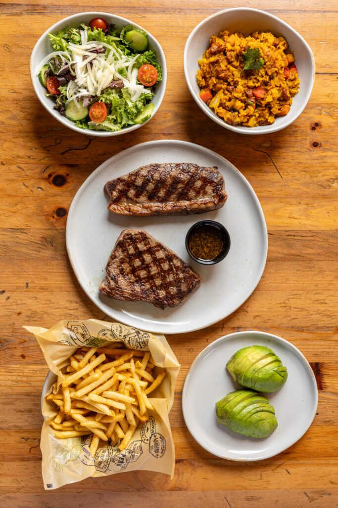 Overhead view of grilled steak cuts served with fries, sliced avocado, mixed green salad, yellow rice, and a small cup of sauce on a wooden table.