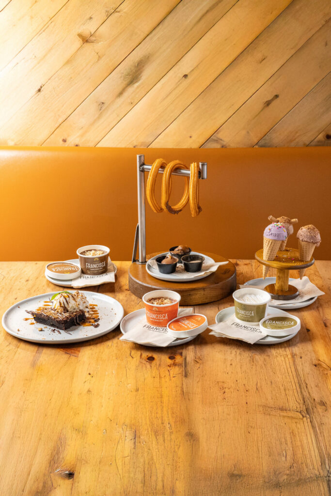 Assorted desserts on a wooden table, including churros with dipping sauces, brownies topped with ice cream, coffee drinks, and ice cream cones displayed on a stand.