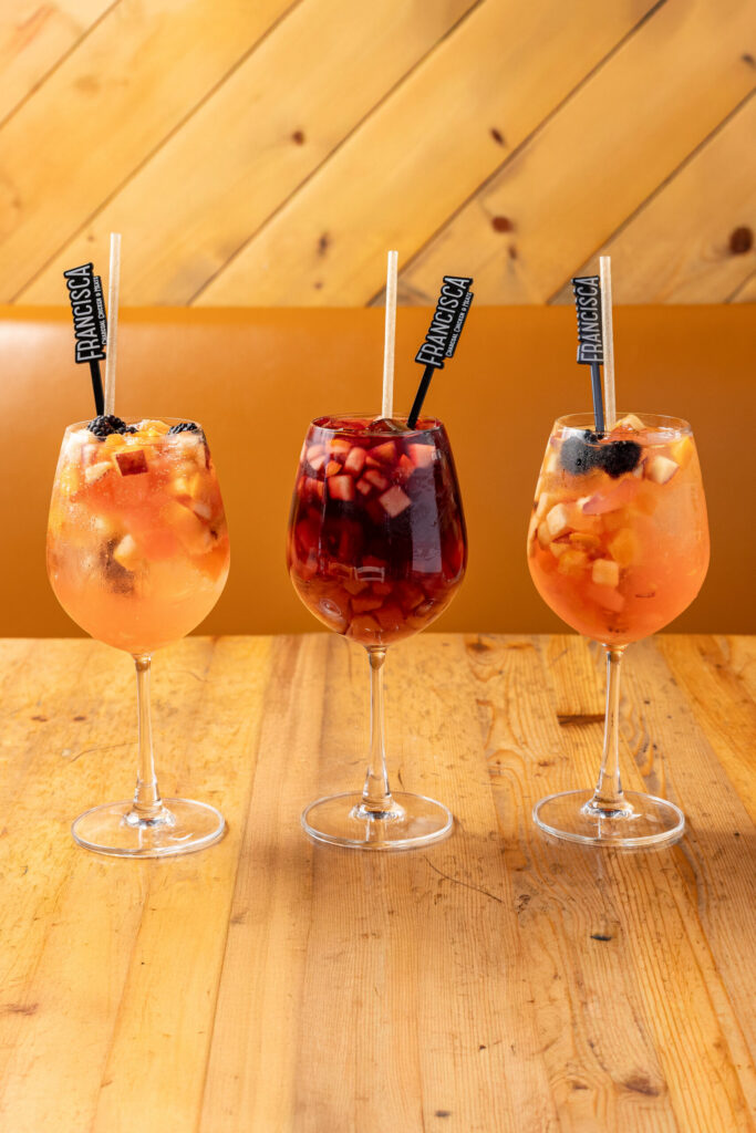 Three fruit sangria cocktails served in wine glasses with ice, chopped fruit, and blackberries on a wooden table.