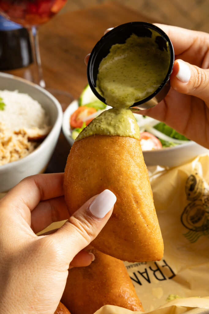Green cilantro sauce being poured over a golden fried empanada held by hand.