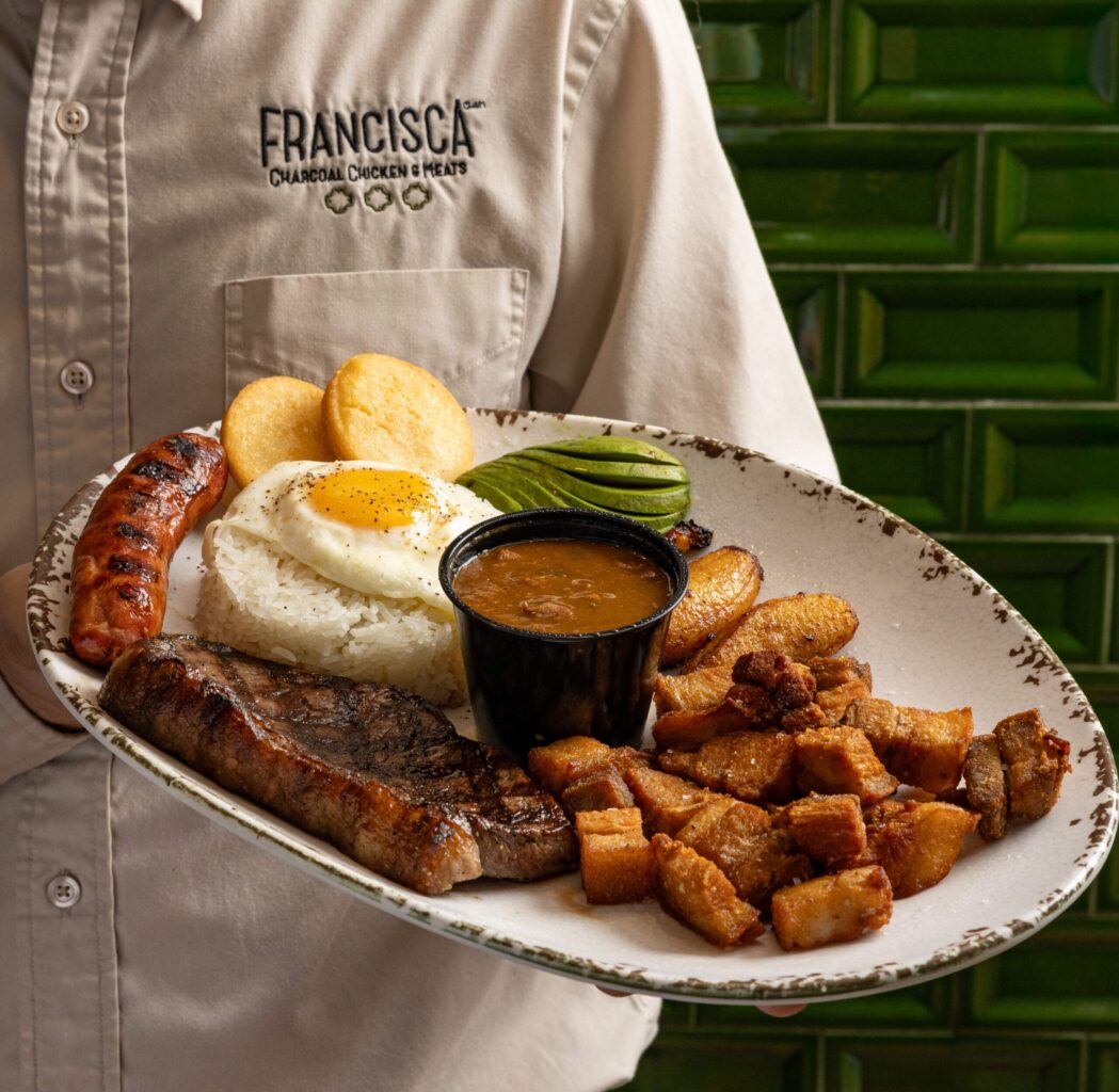 Francisca server holding a large platter with grilled steak, sausage, fried pork belly, rice with fried egg, avocado, plantains, beans, and arepas